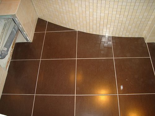 Brązowe, błyszczące płytki podłogowe w łazience, ułożone do krawędzi jasnych, mozaikowych płytek ściennych; widoczny fragment rury kanalizacyjnej w narożniku.